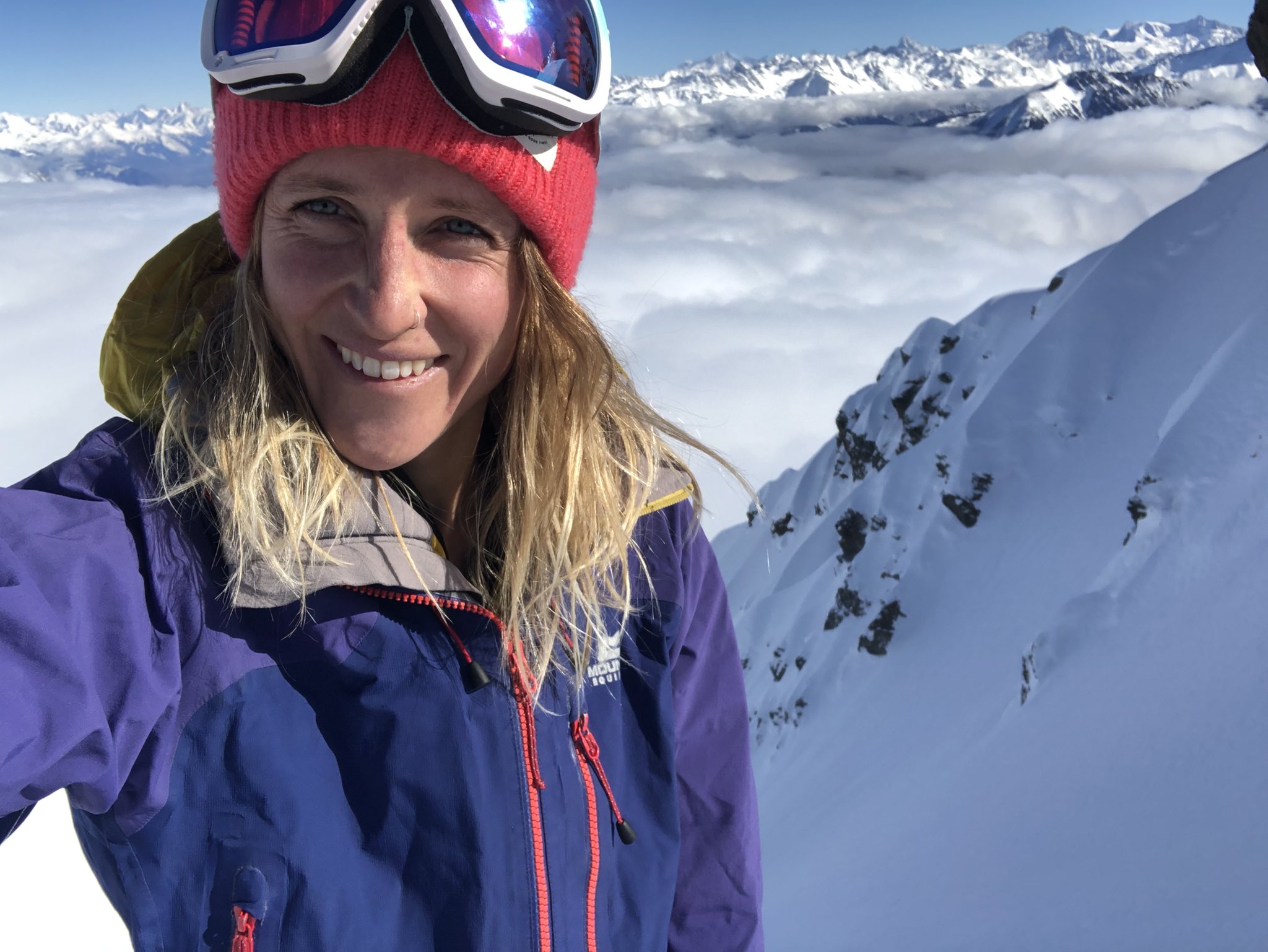 A woman in heavy snow gear atop the clouds in a mountain range.