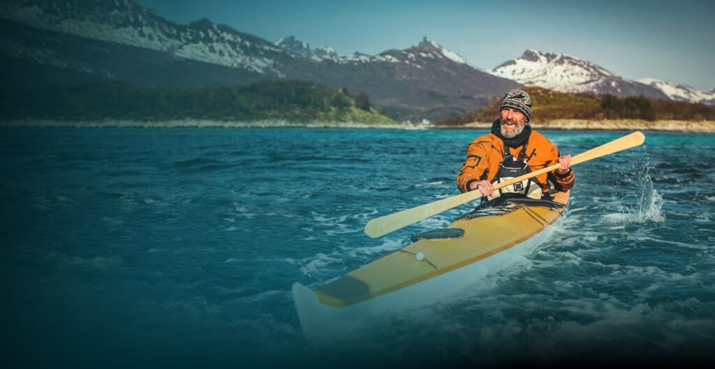 A beared man using a canoe against a snowy mountain range.